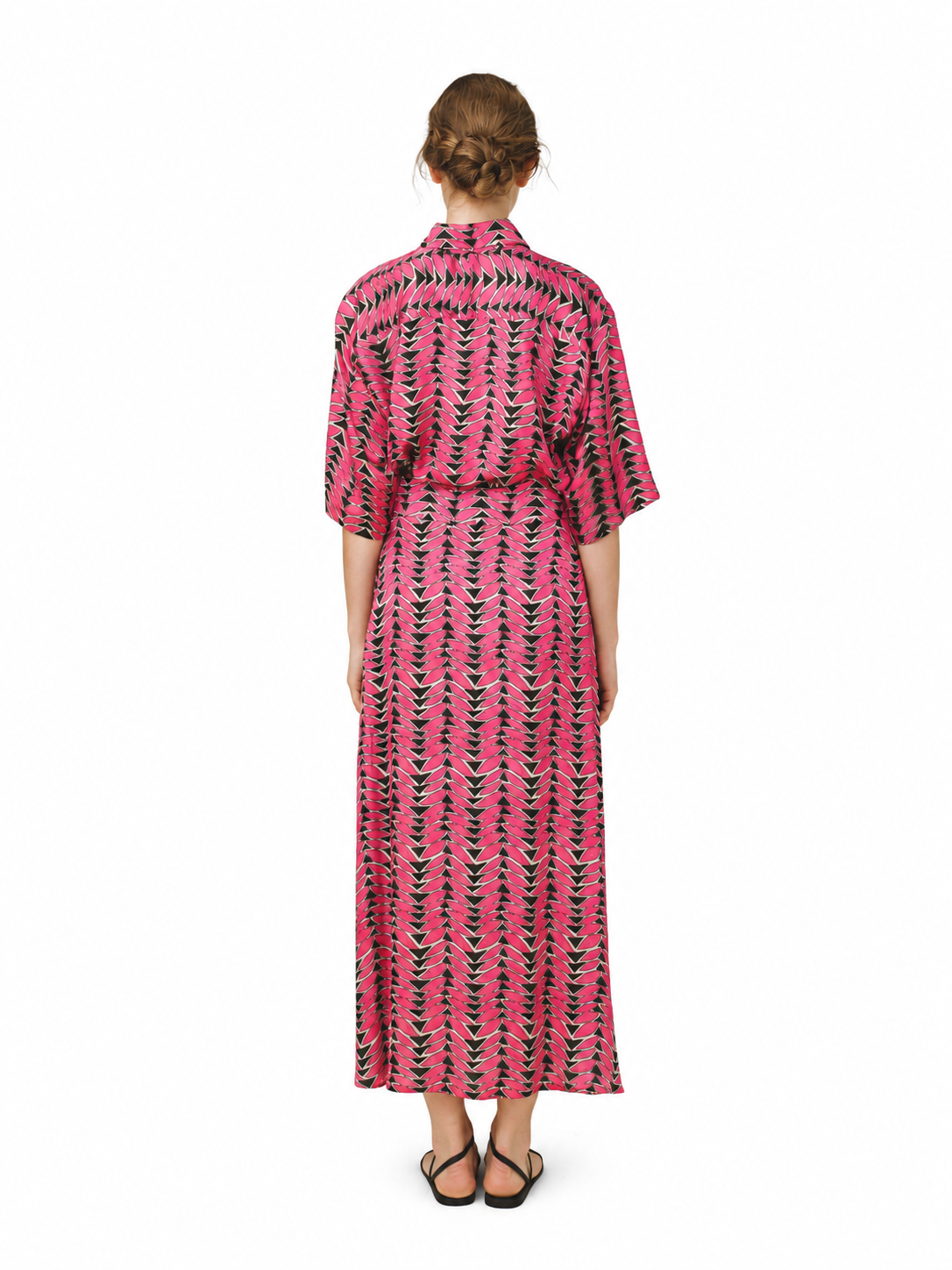 A woman viewed from the back wearing a pink maxi shirt dress with a black and green vertical geometric pattern, set against a plain white background.