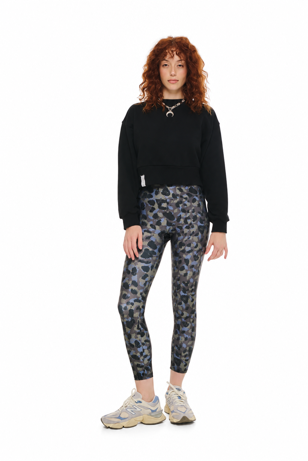 A model with curly red hair stands against a white background wearing a black cropped sweatshirt, blue and gray leopard-print leggings, and beige sneakers.