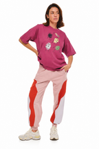 A model wearing a pink graphic t-shirt and light pink jogger pants with red and white side panels, paired with white sneakers, against a solid white background.