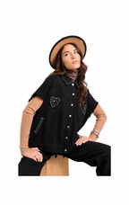 A woman models a black denim vest with studded heart and pocket details, layered over a tan short-sleeve top and black pants, while wearing a tan hat and sitting on a wooden block against a white background.