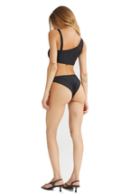 A woman shown from the back wearing a black two-piece bikini with a one-shoulder top and high-cut bottoms, paired with black strappy high-heeled sandals, against a plain white background.