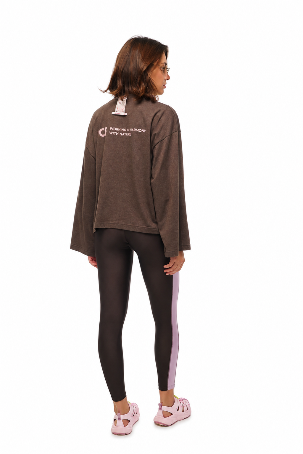 A woman is shown from the back wearing a dark brown long-sleeve sweatshirt with a white printed logo on the back, paired with black leggings featuring a light purple side stripe and pink sneakers, against a plain white background.