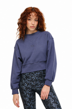A woman with curly red hair wearing a faded slate blue cropped crewneck sweatshirt and black patterned leggings, photographed against a solid white background.