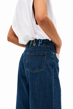 A close-up side view of a person wearing dark blue wide-leg denim jeans with colorful paperclip belt loops (green, blue, gold) and a white sleeveless top, against a plain white background.