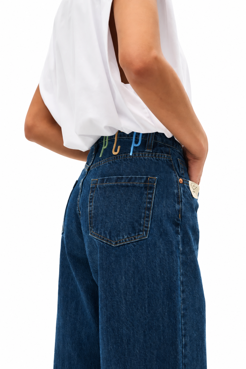 A close-up side view of a person wearing dark blue wide-leg denim jeans with colorful paperclip belt loops (green, blue, gold) and a white sleeveless top, against a plain white background.