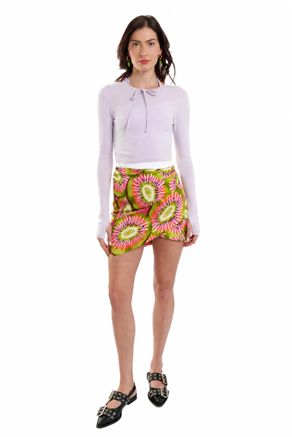 A model stands against a white background wearing a light purple long-sleeve tie-neck top, a vibrant green and pink floral mini skirt, and black studded Mary Jane shoes.