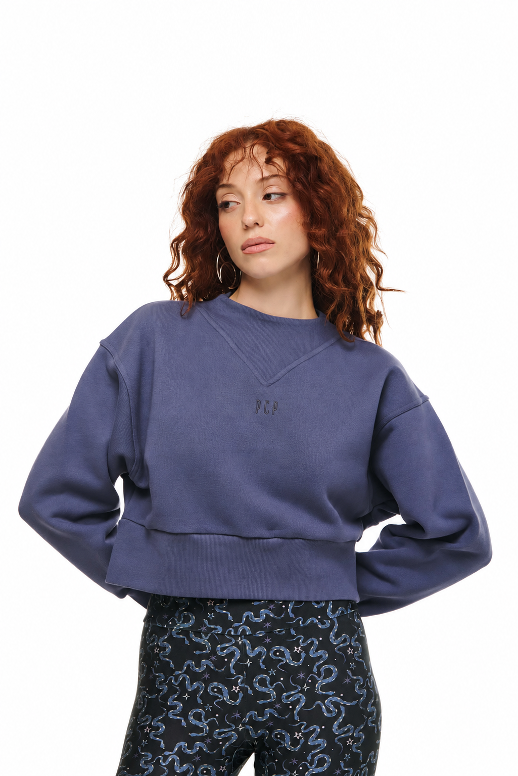 A model with curly red hair wearing a cropped, faded slate blue sweatshirt and patterned leggings, photographed against a solid white background.