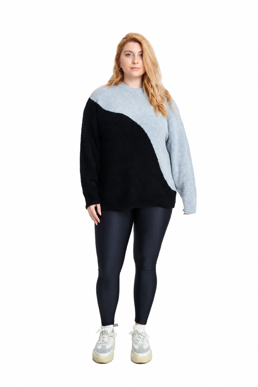 A woman modeling a color-block knit sweater with a black body and a light gray diagonal yoke, paired with dark leggings and white sneakers, on a solid white background.