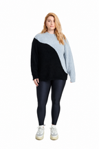 A woman modeling a color-block knit sweater with a black body and a light gray diagonal yoke, paired with dark leggings and white sneakers, on a solid white background.
