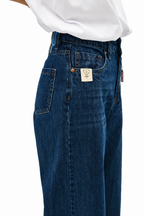 Close-up of a person wearing dark blue denim jeans with a white shirt tucked in, showing the back pocket and a small rectangular brand patch.