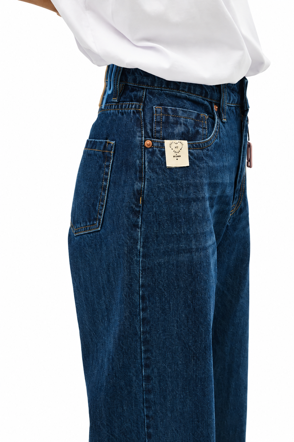 Close-up of a person wearing dark blue denim jeans with a white shirt tucked in, showing the back pocket and a small rectangular brand patch.