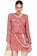 A model wearing a red and white tweed mini skirt suit with pearl accents, including a cropped jacket, matching skirt, and layered pearl chains, holding a pearl handbag against a white background.