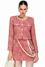 A model wearing a red and white tweed mini skirt suit with pearl accents, including a cropped jacket, matching skirt, and layered pearl chains, holding a pearl handbag against a white background.
