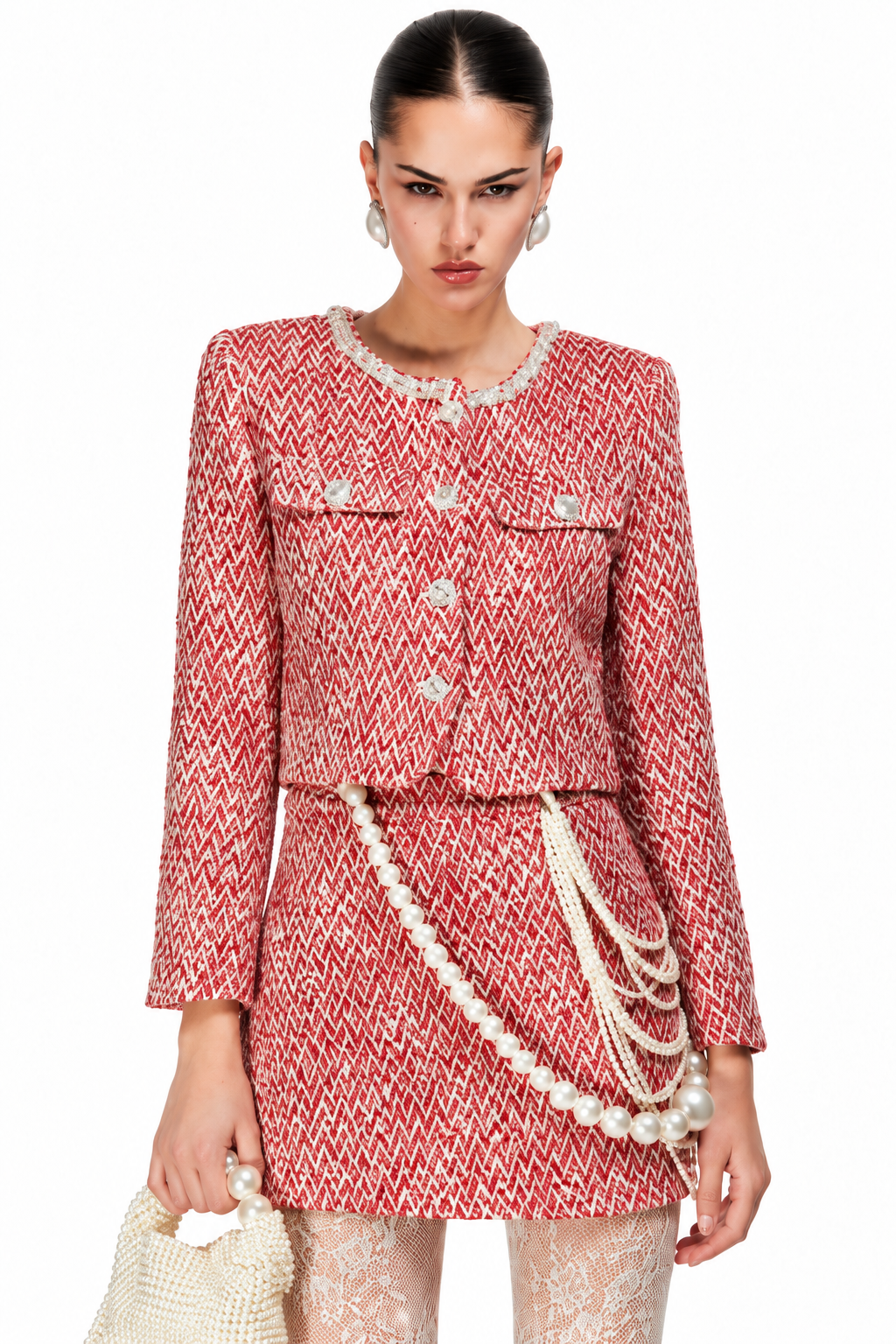 A model wearing a red and white tweed mini skirt suit with pearl accents, including a cropped jacket, matching skirt, and layered pearl chains, holding a pearl handbag against a white background.