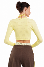 A woman shown from the back wearing a pale yellow long-sleeve crop top with a subtle tonal allover pattern and a tie-neck detail, paired with brown pants against a plain white background.