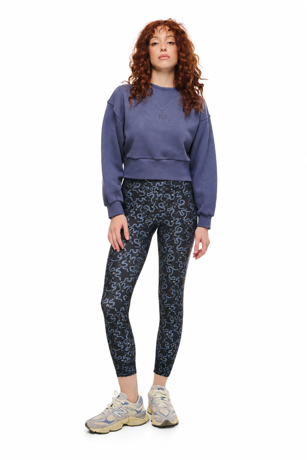 A woman with curly red hair stands against a white background wearing a dusty purple cropped sweatshirt, dark blue patterned leggings, and beige sneakers.