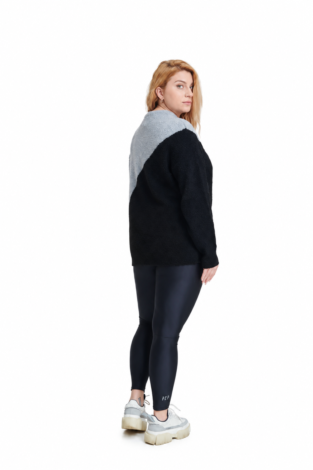 A woman stands in profile wearing a color-block knit sweater with a black body and a heather gray yoke, paired with black leggings and white platform sneakers, against a plain white background.