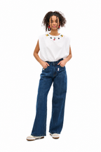 A woman modeling a white sleeveless top with colorful embroidered details around the neckline, paired with high-waisted, wide-leg blue jeans and white sneakers, against a plain white background.