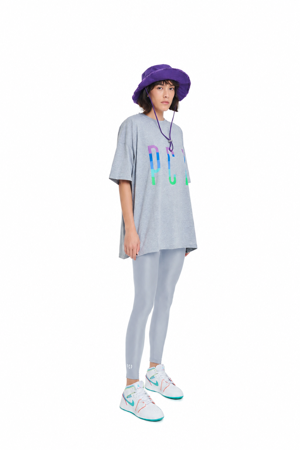A model stands against a white background wearing a light gray oversized t-shirt with a colorful graphic, matching gray leggings, white sneakers with teal accents, and a purple bucket hat.