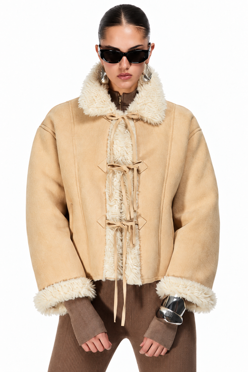 A model wearing a beige shearling-lined jacket with tie-front closures and a high fluffy collar, paired with brown ribbed pants and black sunglasses, against a plain white background.