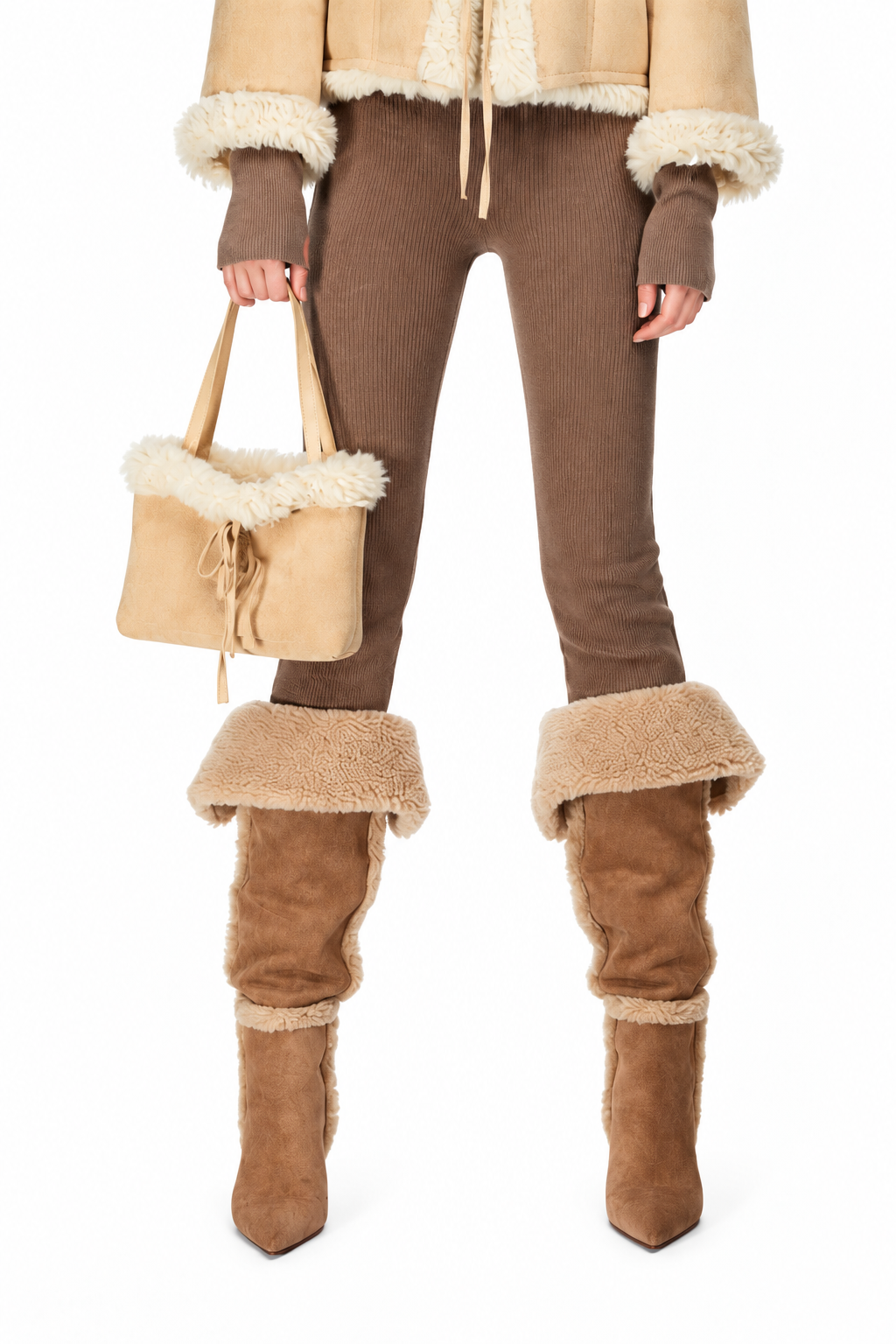 A model is shown from the waist down wearing a matching beige and brown shearling-lined outfit, including a cropped jacket, ribbed brown pants, matching shearling boots, and holding a matching shearling handbag against a white background.