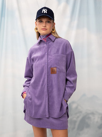 A female model wearing an oversized purple corduroy shirt with a classic collar, front button fastening, chest pocket, and vegan-leather logo patch to the chest. She is also wearing a NY Yankees baseball cap and earrings.