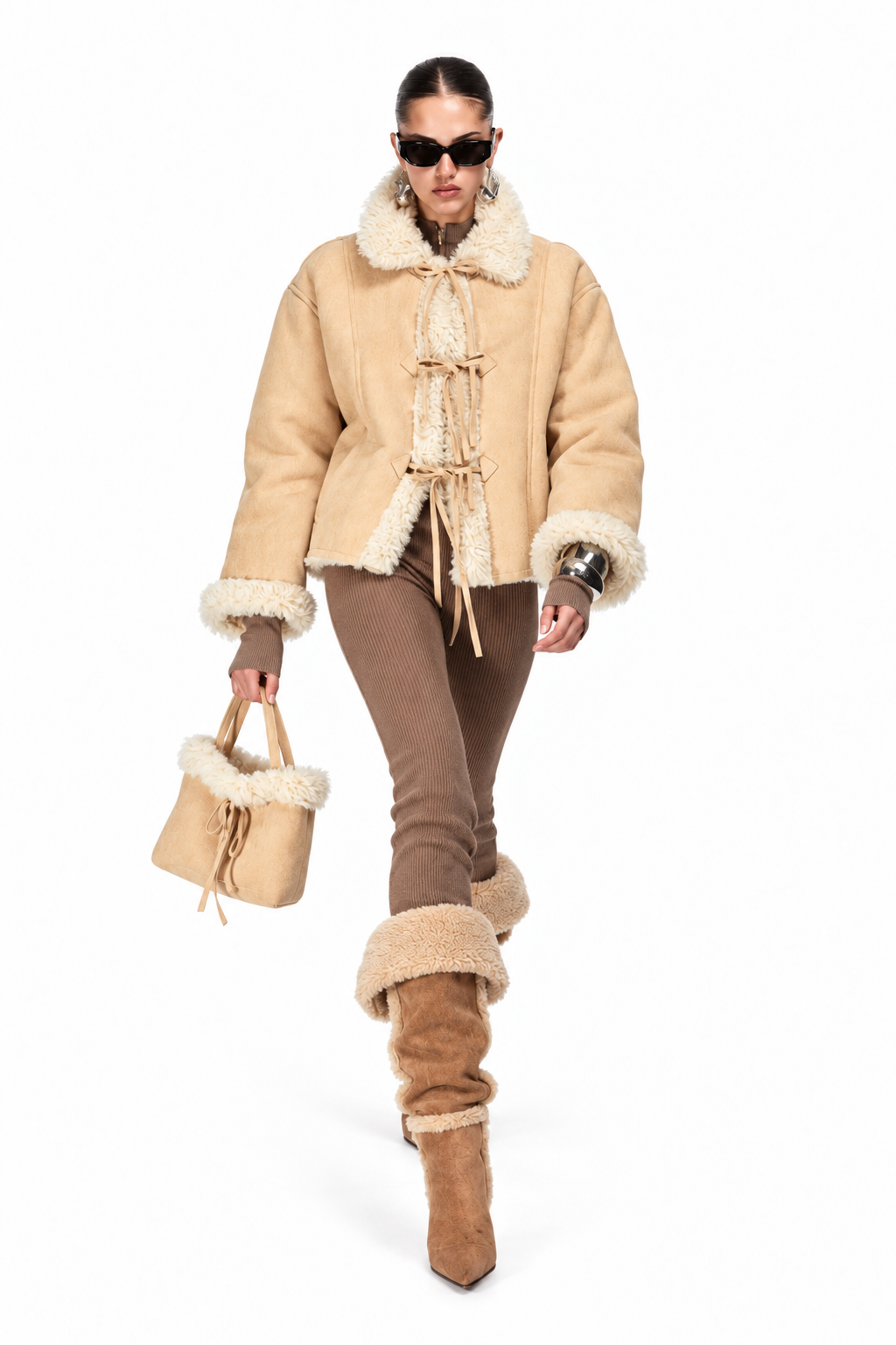 A model walks against a white background wearing a beige shearling jacket with a high collar, brown pants, and brown slouchy boots with shearling trim, holding a matching beige shearling handbag.
