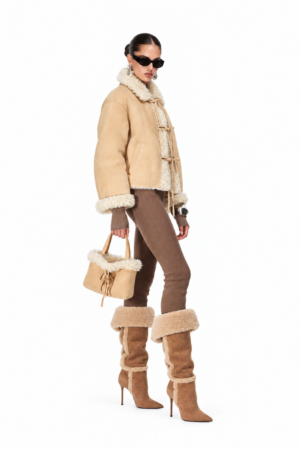 A model wearing a beige shearling-lined jacket with toggle closures, brown pants, and matching shearling-trimmed boots, holding a matching beige and cream handbag against a plain white background.