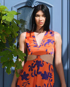 Woman wearing an orange and blue floral dress standing against a blue wall with green leaves.
