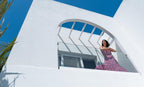 Woman in a pink dress standing on a white balcony with blue sky and palm trees.