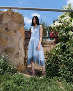 Woman in a light blue dress standing outdoors near a stone wall and flowers.