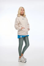 Woman wearing a beige hoodie with a logo, blue skirt, green leggings, and white sneakers on a white background