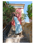 Woman in a light blue dress standing on stone steps with greenery and a building in the background