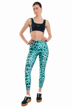 A woman modeling a matching activewear set featuring a black sports bra with teal and black leopard print trim, and high-waisted teal leggings with a black leopard print pattern. She is wearing black sneakers with white stripes and brown soles, posed against a solid white background.