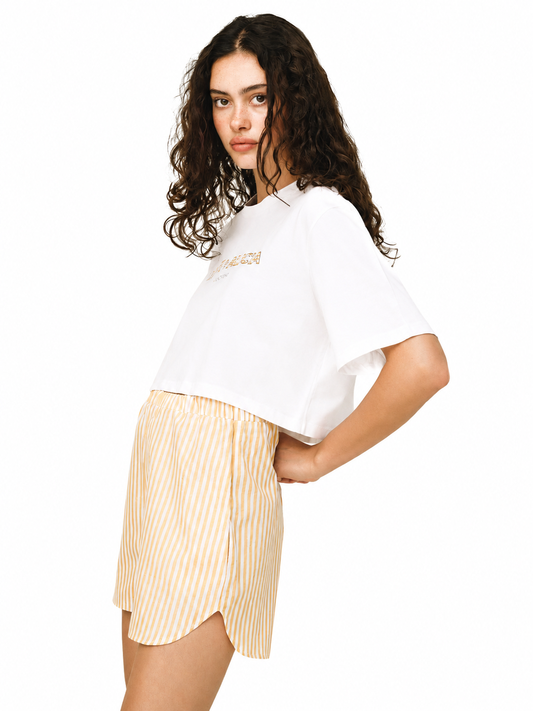 A model wearing a white cropped t-shirt with gold lettering and yellow-and-white vertically striped shorts, photographed against a solid white background.