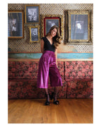 Woman in a black top and pink pants standing in a room with floral wallpaper and framed pictures.