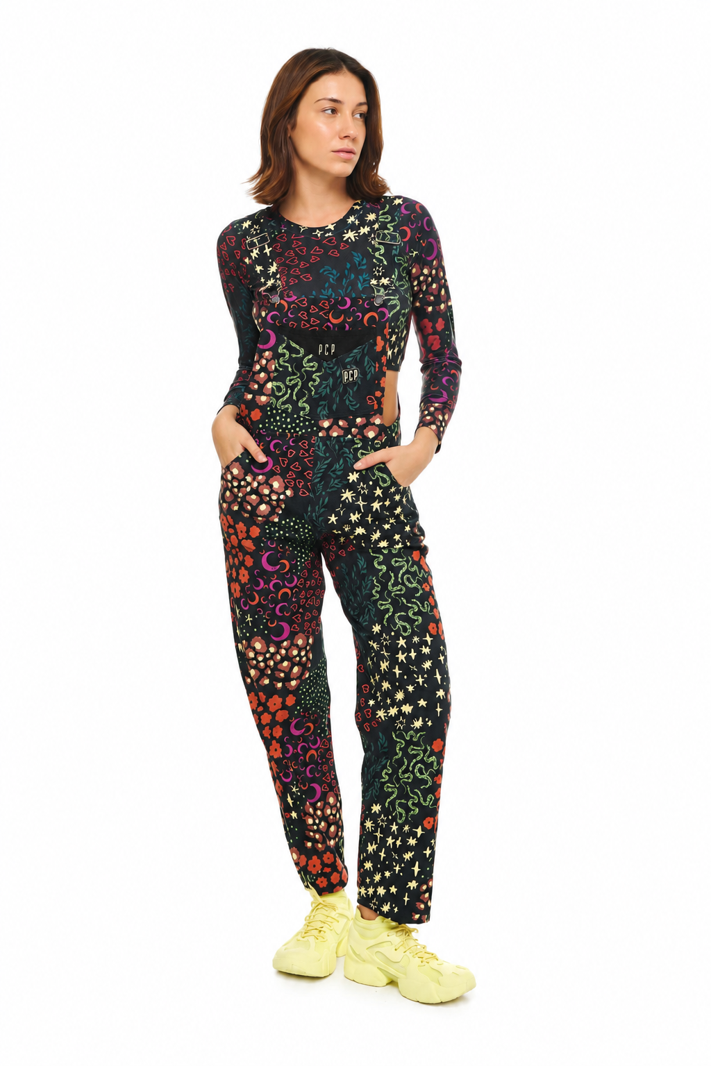 A woman modeling a matching long-sleeve crop top and jogger pants set with a vibrant multicolor bohemian floral pattern on a black background, styled with bright yellow sneakers against a plain white studio background.