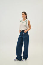 Woman wearing a white tank top with green trim and blue jeans on a light gray background