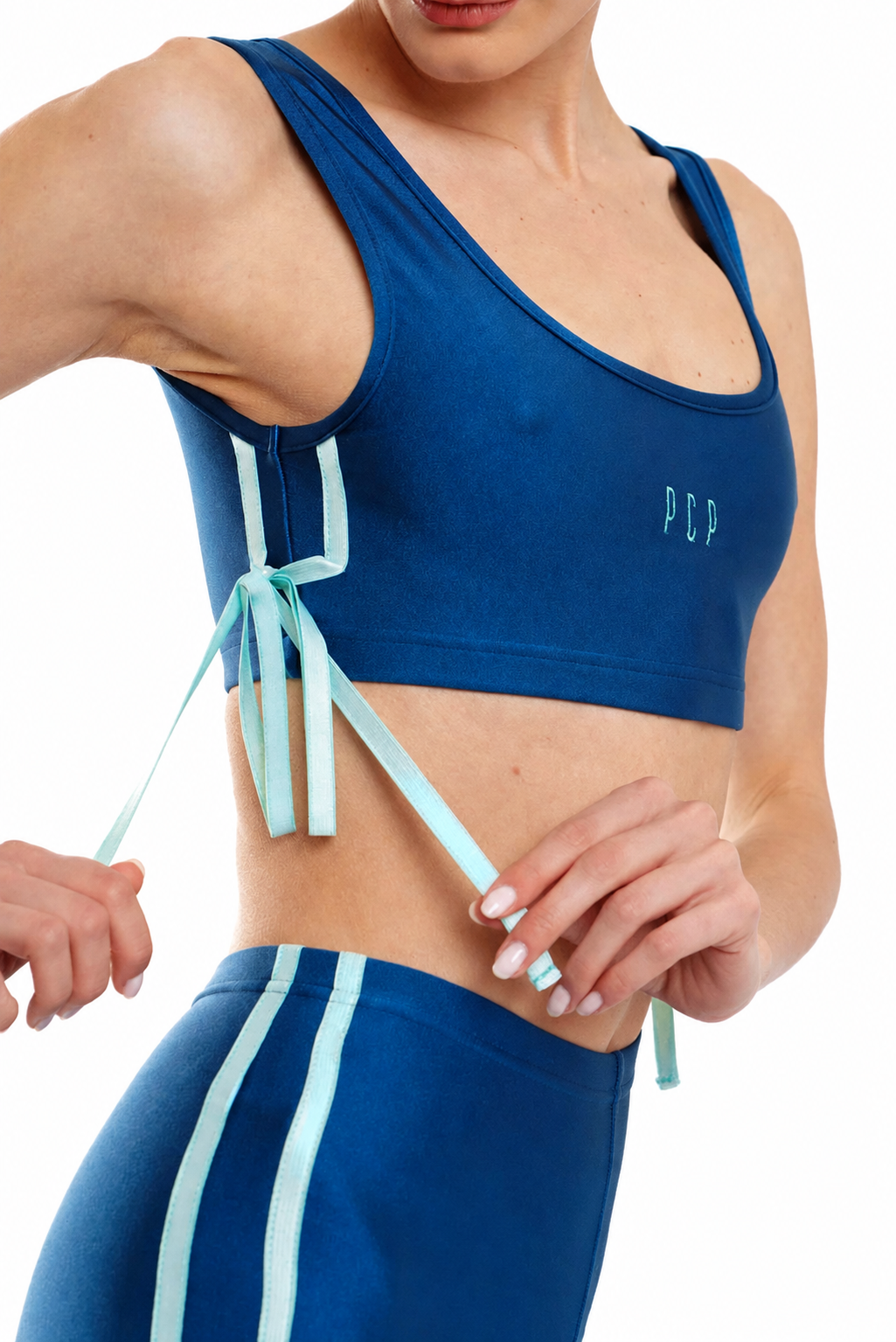 A close-up of a woman wearing a royal blue athletic sports bra with mint green tie-front laces and matching blue leggings with white side stripes. The brand name 'PDP' is embroidered on the chest.