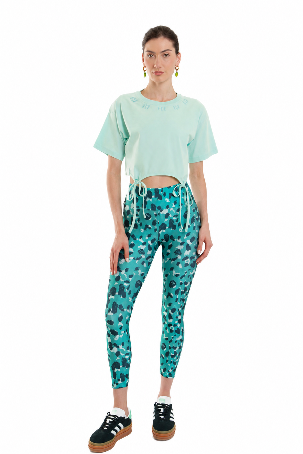 A model stands against a white background wearing a mint green cropped t-shirt, teal and black patterned leggings, and black platform sneakers.
