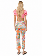 A woman viewed from behind wearing a bright pink puff-sleeve crop top and high-waisted cropped pants with a colorful tropical leaf print in orange, teal, pink, and white, paired with black sandals on a white background.