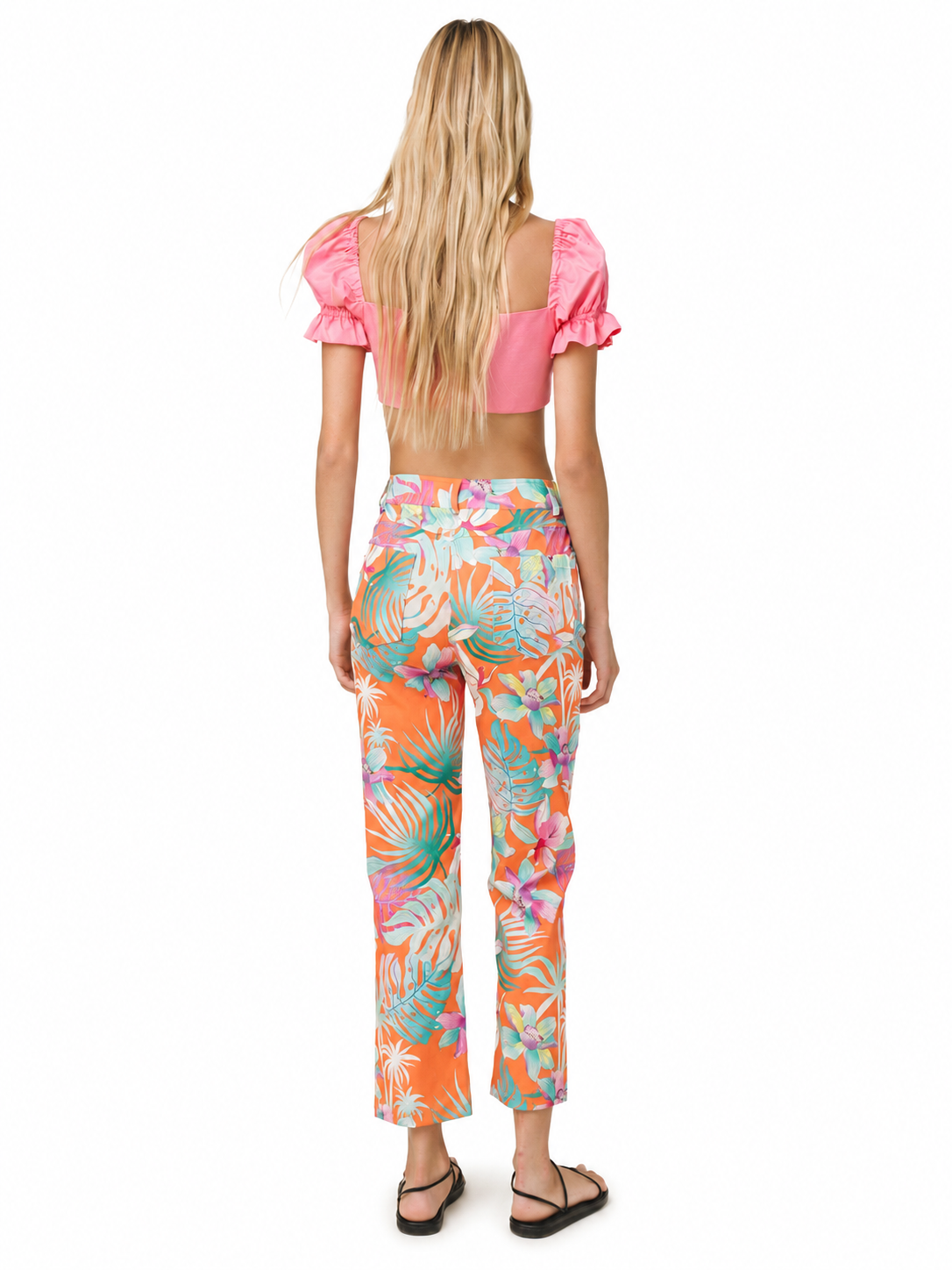 A woman viewed from behind wearing a bright pink puff-sleeve crop top and high-waisted cropped pants with a colorful tropical leaf print in orange, teal, pink, and white, paired with black sandals on a white background.