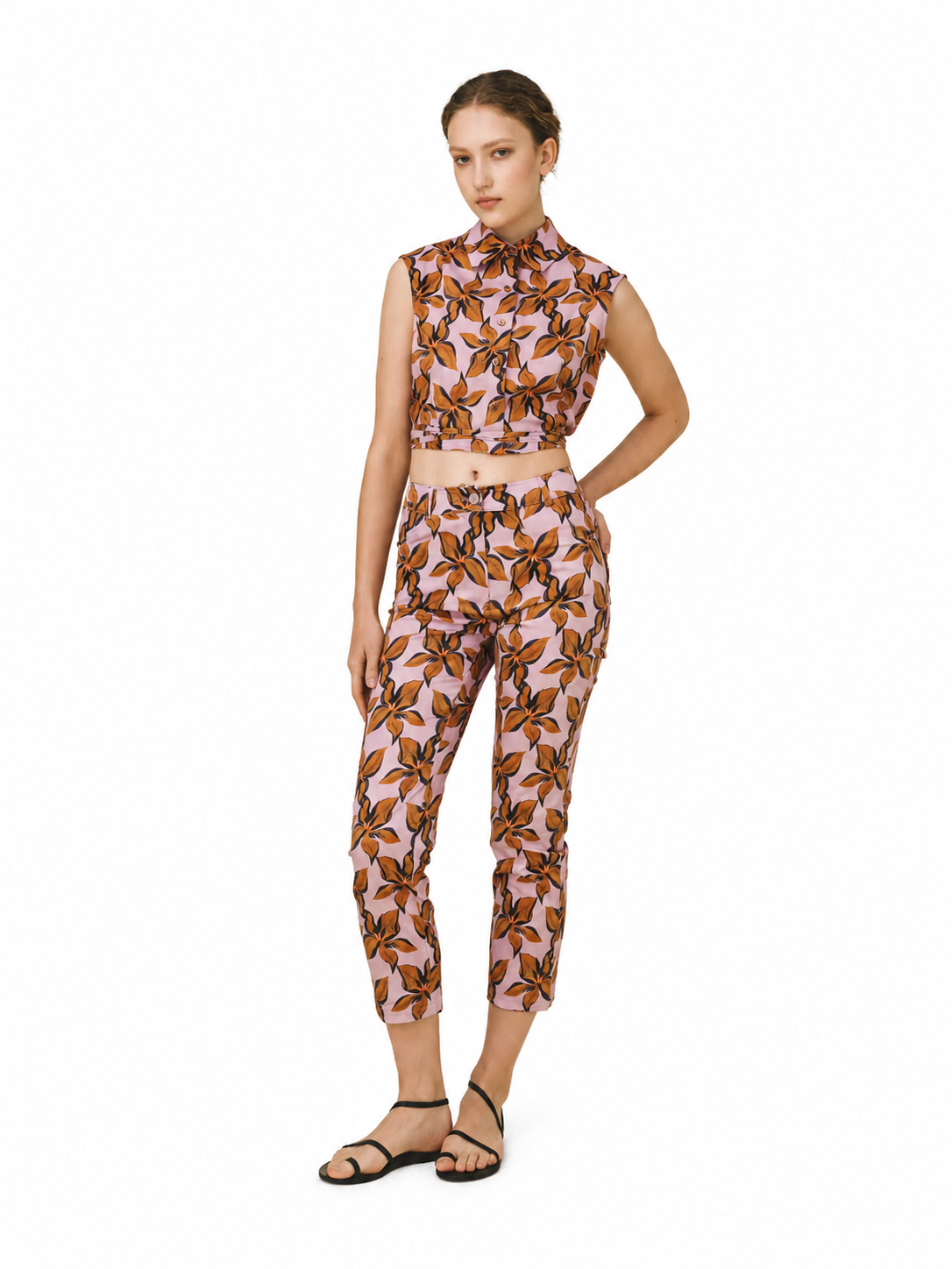 A woman modeling a matching sleeveless crop top and cropped pants set with a pink, brown, and orange botanical leaf pattern, paired with black strappy flat sandals, on a solid white background.