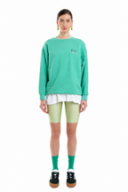 A model stands against a white background wearing a mint green crewneck sweatshirt with a small white graphic on the left chest, styled with a white undershirt, light green biker shorts, green socks, and black platform sneakers.