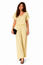 A woman modeling a pale yellow short-sleeve jumpsuit with a V-neckline and wide-leg pants, holding a small black clutch against a plain white background.