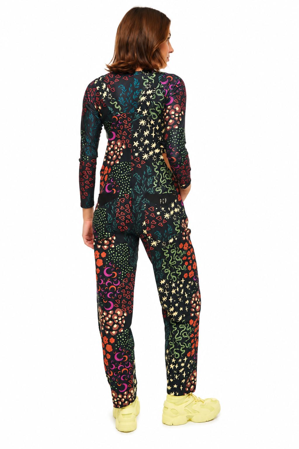 A woman viewed from the back wearing a long-sleeve one-piece jumpsuit with a dense multicolor abstract floral and dot pattern on a black base, paired with light yellow sneakers, against a plain white background.