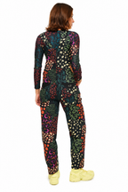 A woman viewed from the back wearing a long-sleeve one-piece jumpsuit with a dense multicolor abstract floral and dot pattern on a black base, paired with light yellow sneakers, against a plain white background.