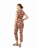 A woman modeling a sleeveless high-neck jumpsuit with a brown and orange leaf pattern on a light pink background, shown from the back against a white studio background.