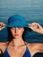 Woman wearing a blue bucket hat with 'Sun of a Beach' text against a blurred ocean background