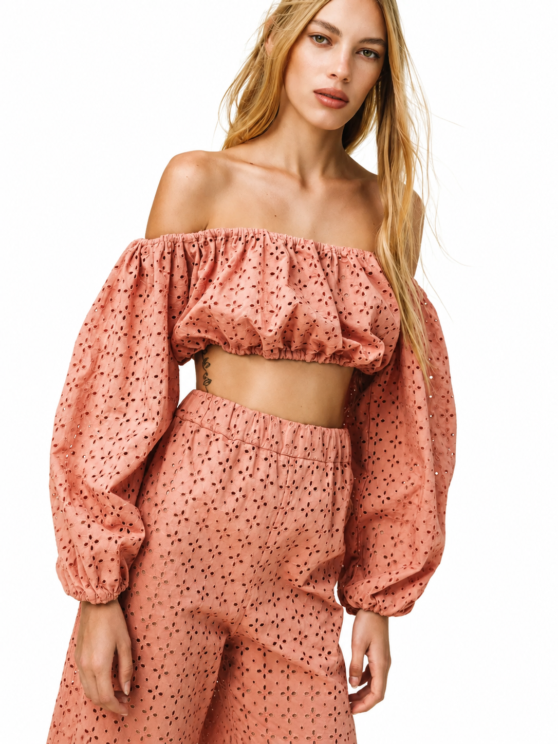 A woman modeling a matching two-piece set featuring an off-the-shoulder crop top and high-waisted trousers, both in a dusty rose pink color with a small black polka dot pattern, set against a plain white background.